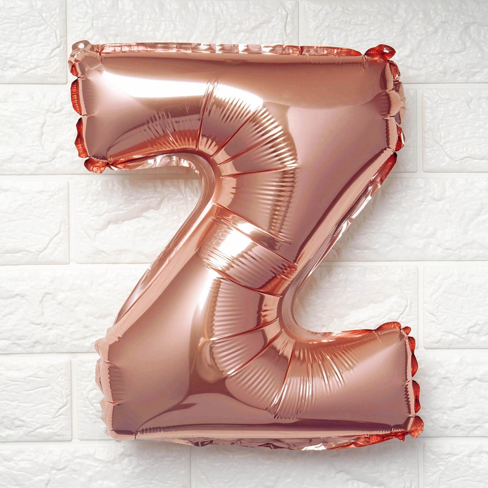 A metallic rose gold foil balloon shaped like the letter 'Z', floating against a white textured brick wall background.