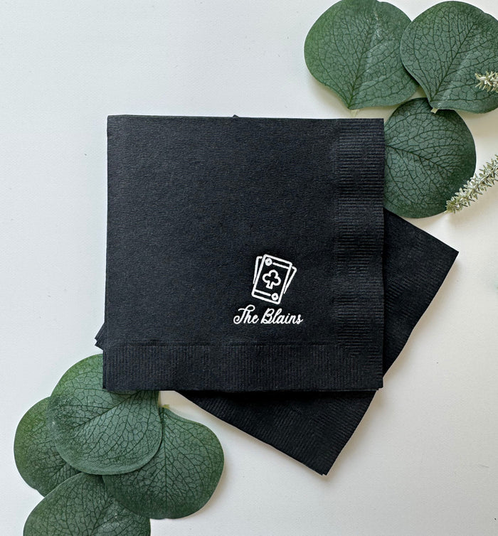 Two black paper napkins with white printed 'The Blanks' logo with playing card motif, arranged on a white surface with decorative green leaves.