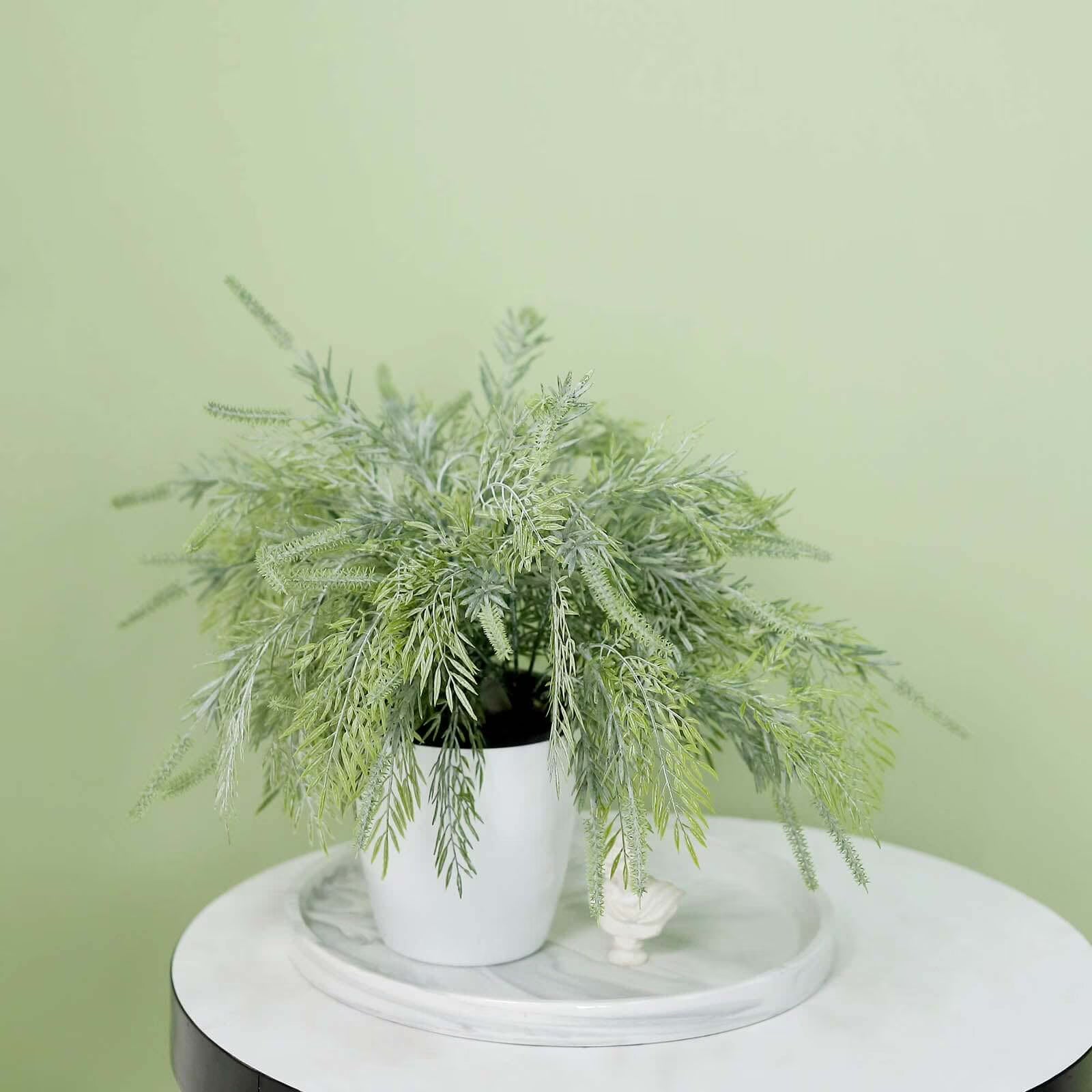 An artificial green fern-like plant in a white ceramic pot, displayed on a round white marble tray atop a white table against a pale green wall.