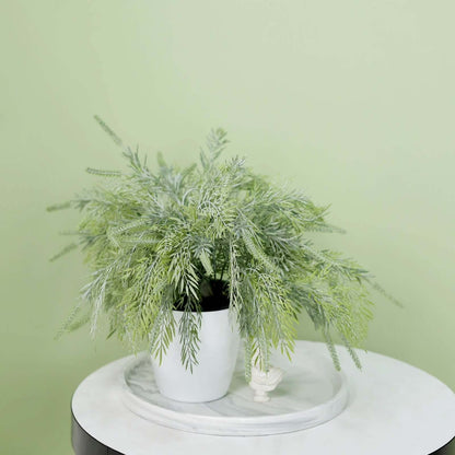 An artificial green fern-like plant in a white ceramic pot, displayed on a round white marble tray atop a white table against a pale green wall.