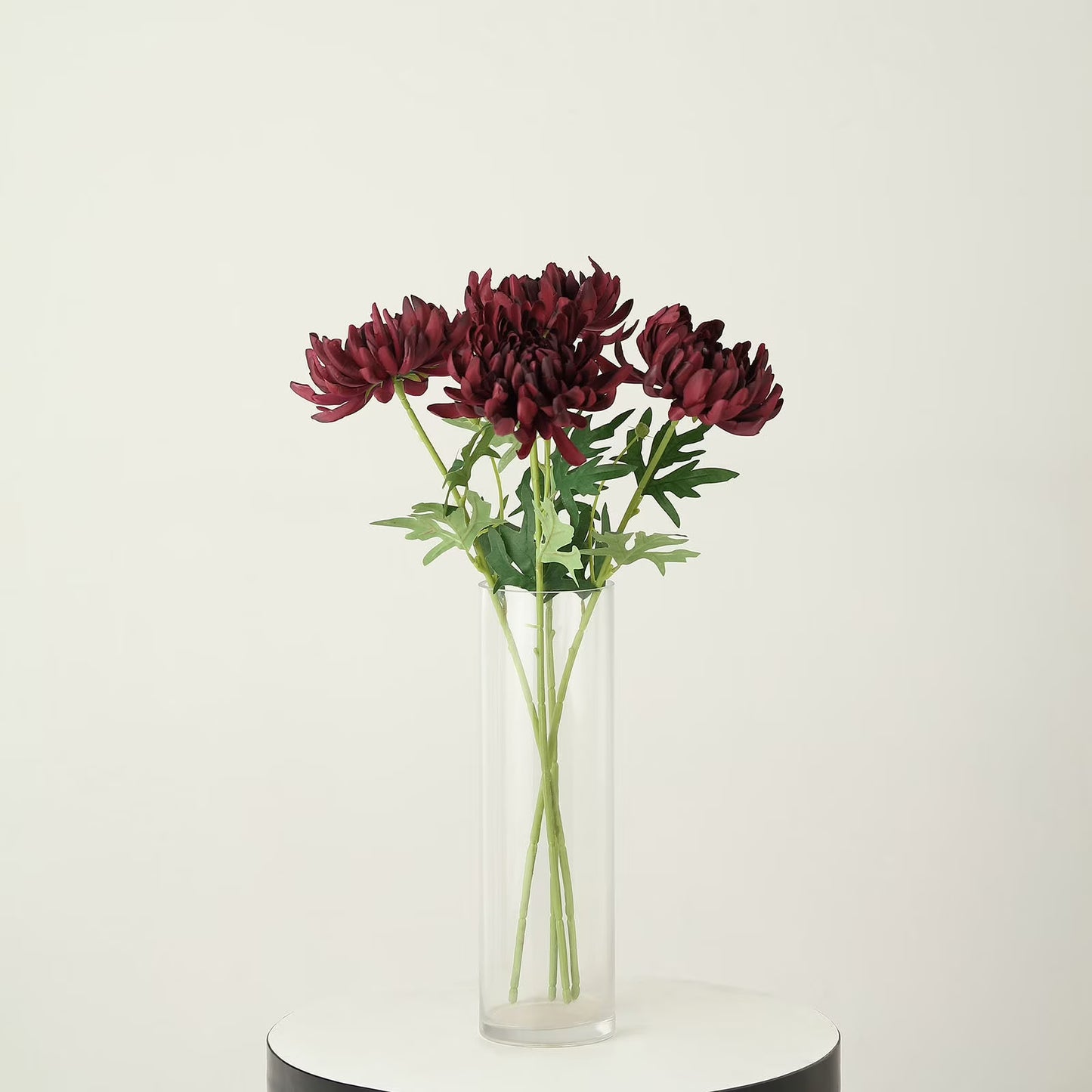 A bouquet of deep burgundy chrysanthemums with green stems and leaves arranged in a clear cylindrical glass vase, set on a round white and black pedestal against a plain off-white background.