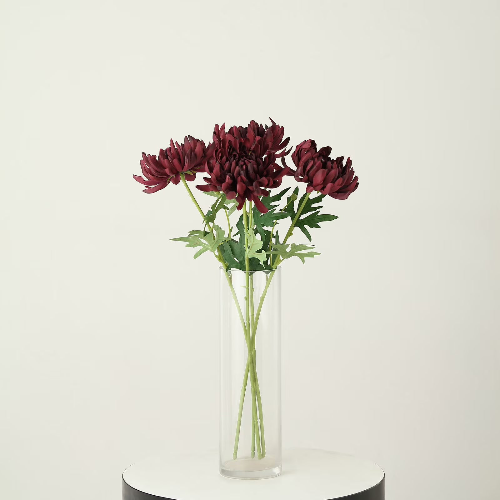 A bouquet of deep burgundy chrysanthemums with green stems and leaves arranged in a clear cylindrical glass vase, set on a round white and black pedestal against a plain off-white background.