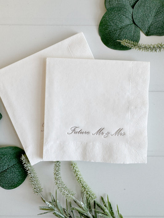 Two personalized white paper napkins stacked on a white wooden surface, with the front napkin embroidered 'Future Mr. & Mrs. for an engagement party or wedding weekend welcome toast.