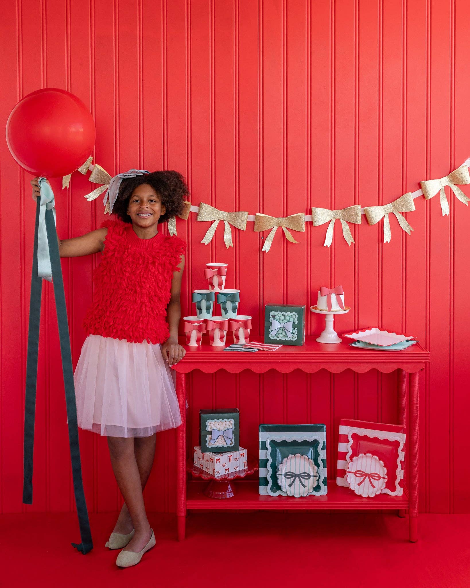 Young girl in a red top and white skirt standing next to a red table with party decorations against a red wall with our gold bow garland.