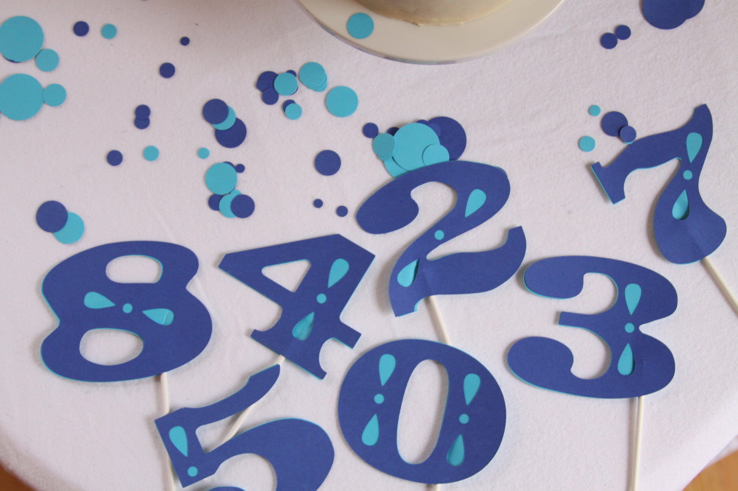 Blue and turquoise number cake toppers on a white frosted cake, including the numbers 8, 4, 2, 3, 1, 5, 0, and 7, with scattered blue and turquoise confetti.