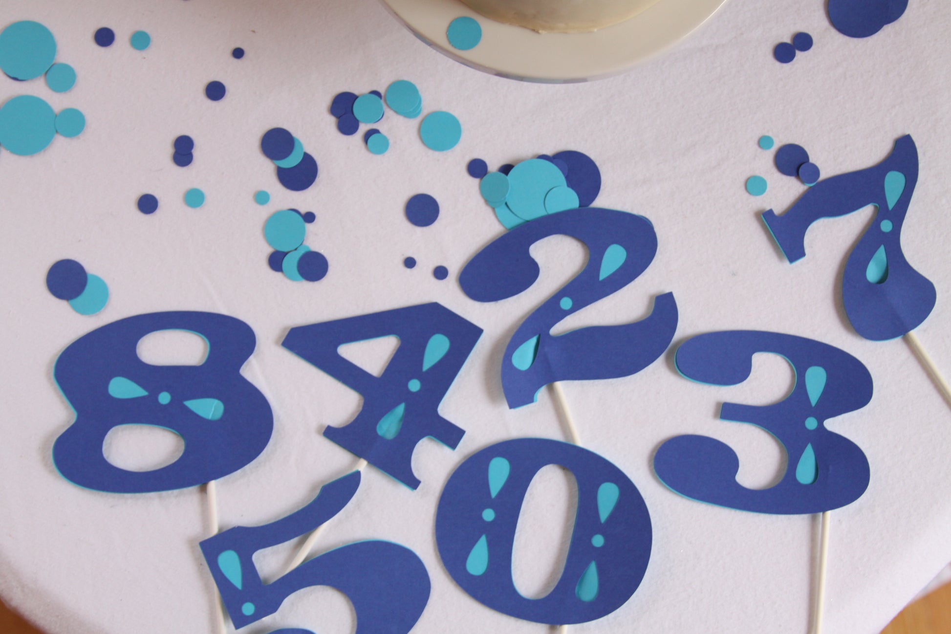 Blue and turquoise number cake toppers on a white frosted cake, including the numbers 8, 4, 2, 3, 1, 5, 0, and 7, with scattered blue and turquoise confetti.