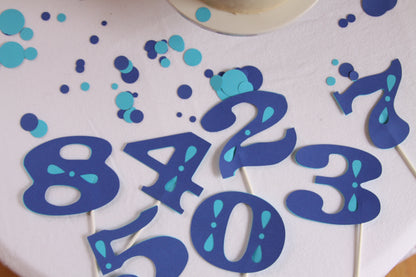 Blue and turquoise number cake toppers on a white frosted cake, including the numbers 8, 4, 2, 3, 1, 5, 0, and 7, with scattered blue and turquoise confetti.