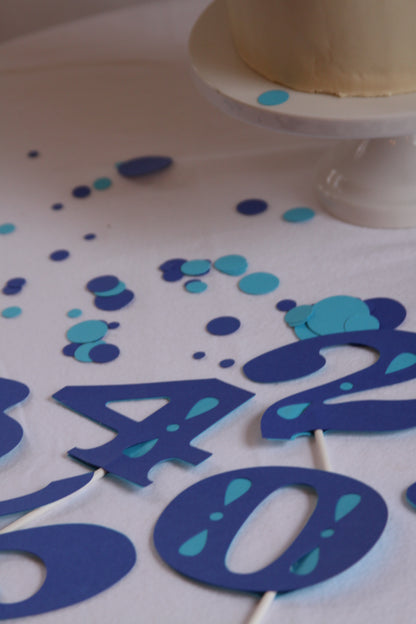 Blue and teal number cake toppers on a white tablecloth, with scattered matching confetti dots and part of a beige cake on a white stand visible in the upper right.