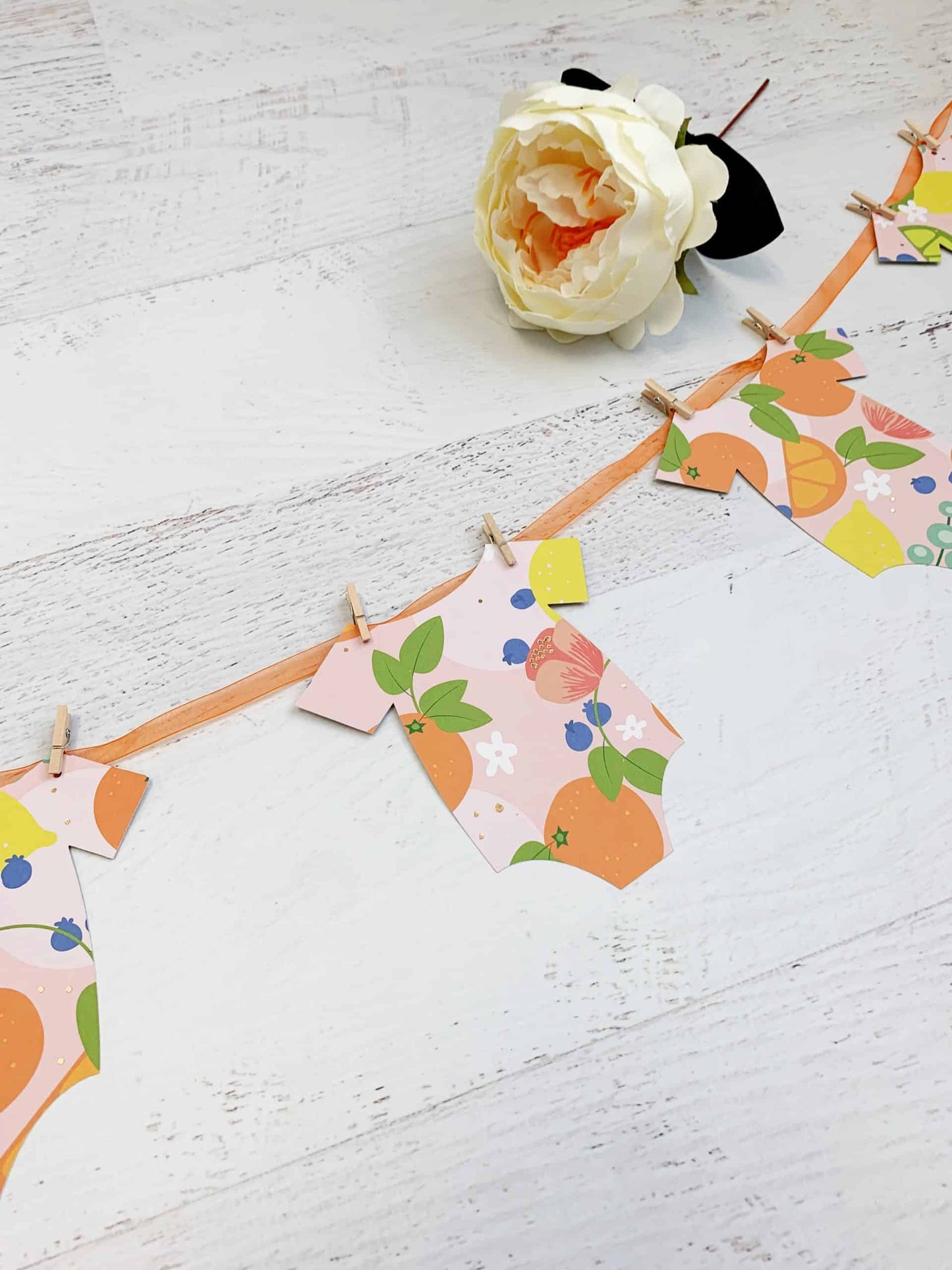 A Little Cutie Citrus Onesie Banner featuring seven onesie-shaped cutouts in bright citrus fruit designs on a blush background, with an orange sheer organza cord and mini clothespins. Perfect for a Little Cutie-themed baby shower.