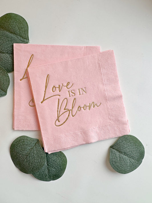 Pink napkins with 'Love is in Bloom' text on a white background with green leaves.