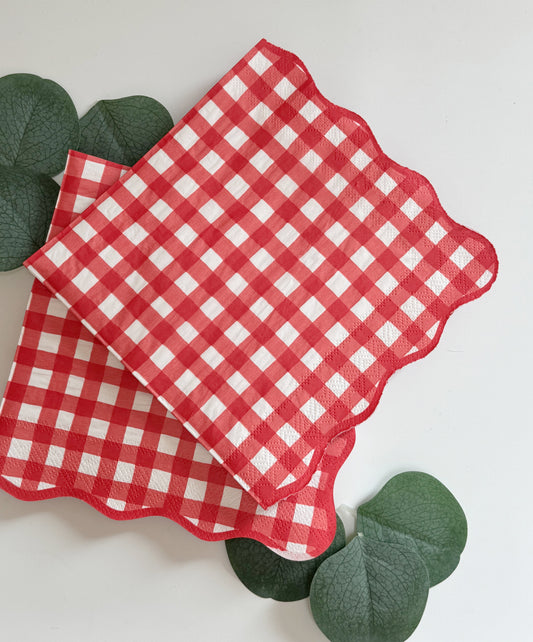 Two red and white gingham checkered paper napkins with scalloped edges, styled with green eucalyptus leaves on a clean white background.