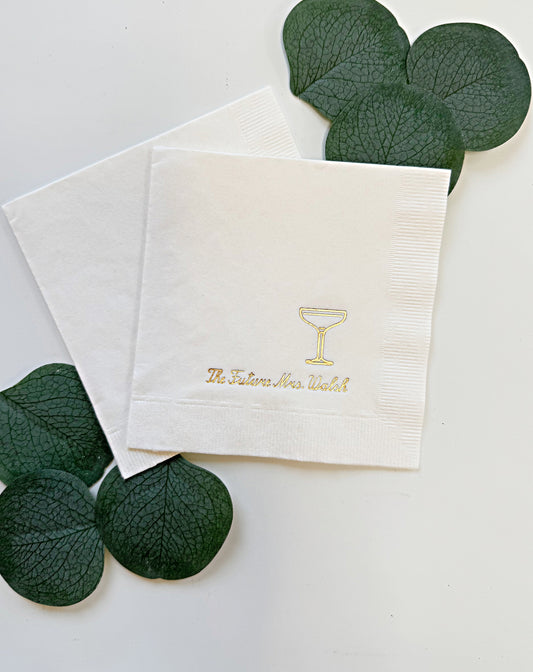Two white personalized paper drink napkins on a white background, one printed with a gold cocktail glass icon and the text 'The Future Ares Watch' in gold. Dark green decorative leaves are arranged around the napkins.