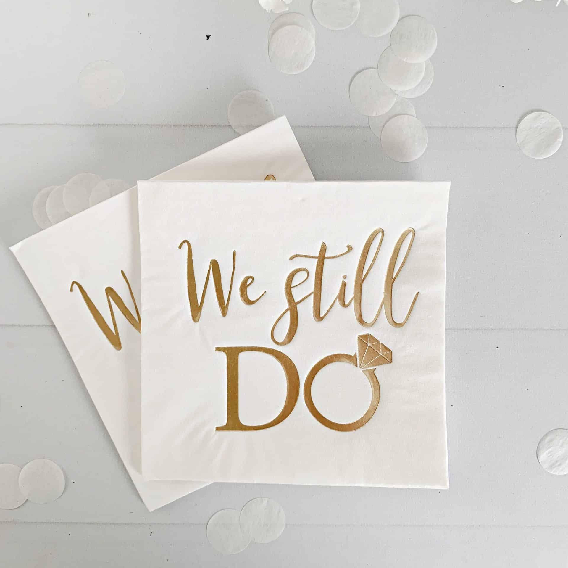 Cocktail napkin with 'We still do' text and a ring symbol on a white background with flowers.