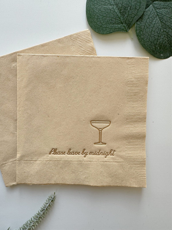 Beige cocktail napkin with gold text  "please leave by midnight" and a martini glass symbol on a white background with green leaves.
