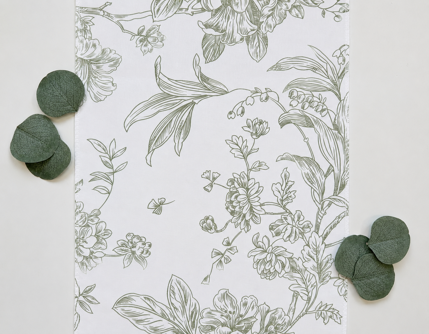 A flat-lay view of a white table runner featuring a delicate sage green botanical line drawing, styled with scattered green eucalyptus leaves on a light neutral background.