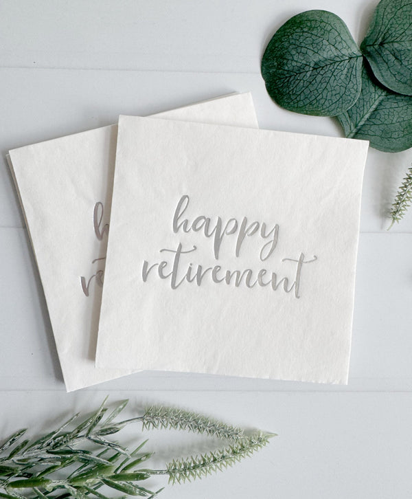 Two white napkins with 'happy retirement' text on a light gray background with green leaves.