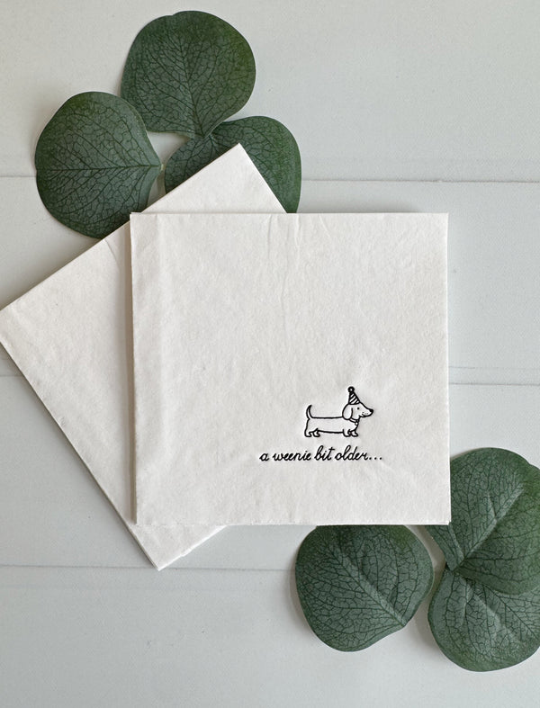 A square white paper birthday napkin stack with a small black line drawing of a dachshund and the text 'a weenie bit older.