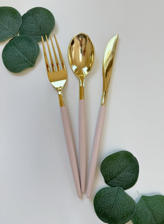 Gold and blush pink cutlery set with a fork, spoon, and knife, styled with green eucalyptus leaves on a white background.