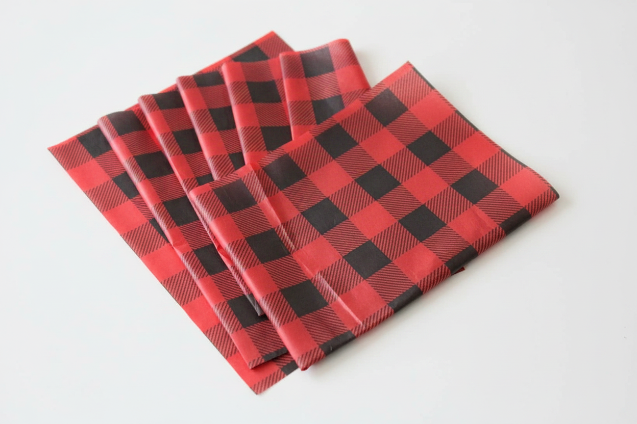 Four folded sheets of tissue paper with a red and black buffalo plaid pattern, arranged in a fan shape on a plain white background.