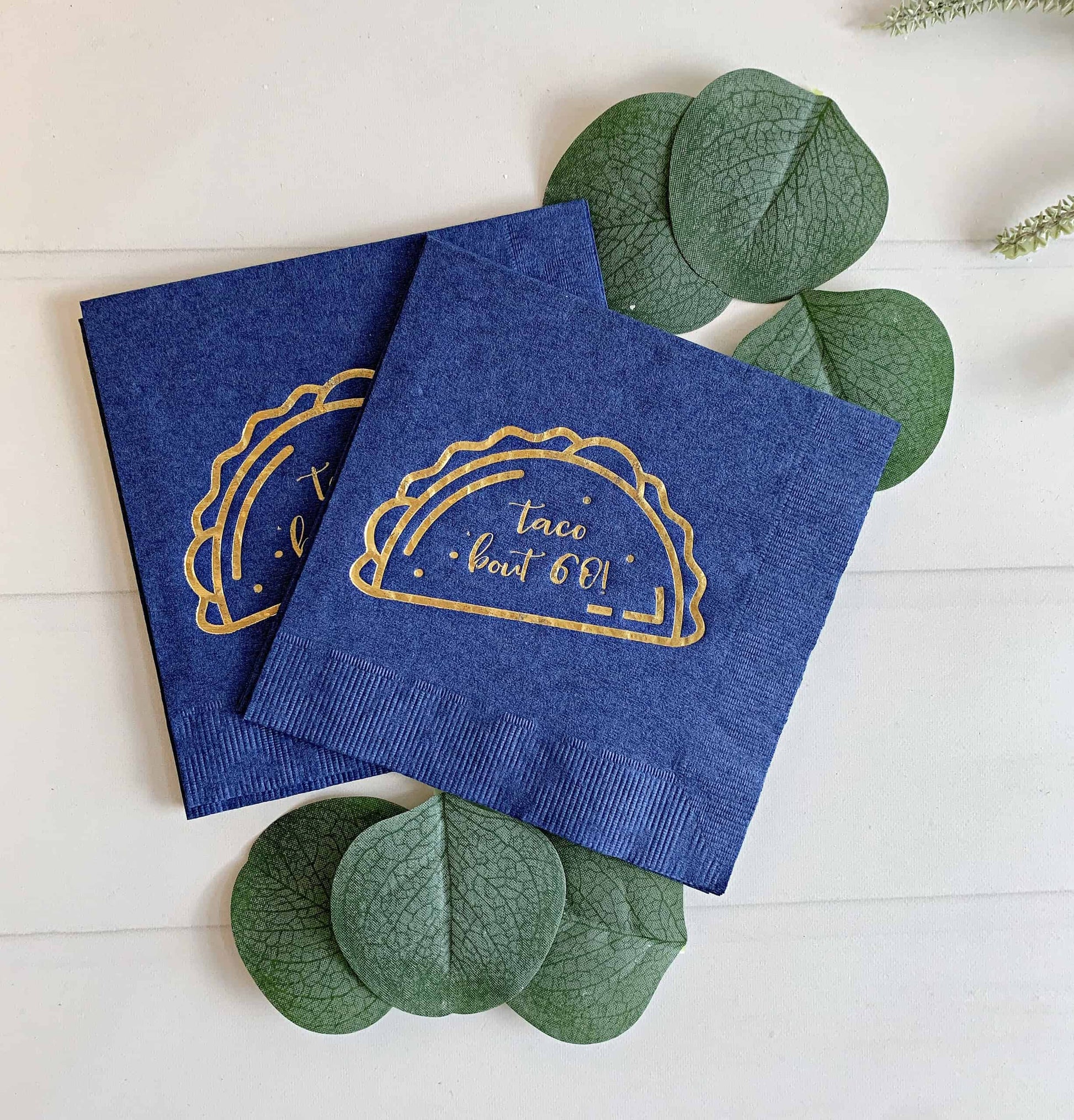 Each cocktail-sized napkin is navy blue and features a gold foil stamp that reads "Taco 'Bout 60." These napkins are perfect for a themed 60th birthday party, especially for a taco bar or fiesta celebration. 