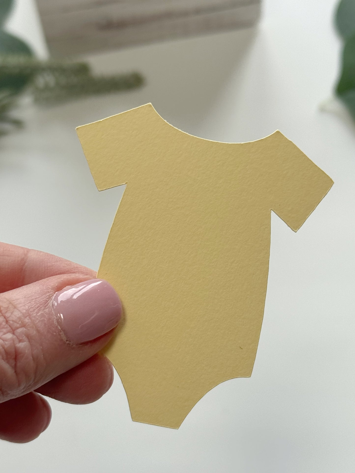 A hand with pink manicured nails holds a small, pale yellow paper cutout shaped like a short-sleeve baby bodysuit against a bright, softly blurred white background.