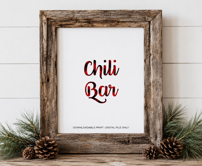 A rustic wooden framed print with the words 'Chili Bar' in red and black plaid-style lettering on a white background. The frame is styled on a wooden surface with pine branches, pinecones, and a white shiplap wall behind it.
