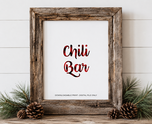 A rustic wooden framed print with the words 'Chili Bar' in red and black plaid-style lettering on a white background. The frame is styled on a wooden surface with pine branches, pinecones, and a white shiplap wall behind it.