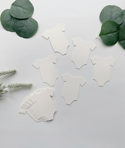 White baby bodysuit-shaped party cutouts arranged on a light gray background with green eucalyptus leaves and small white flowers as decorative props.