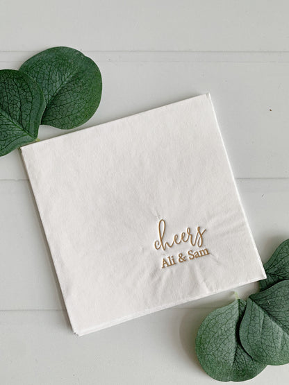 A white paper coktail napkin with gold foil text reading 'cheers' and  personalized with 'Ali & Sam' in a cursive font, styled on a white wood background with green leaves.