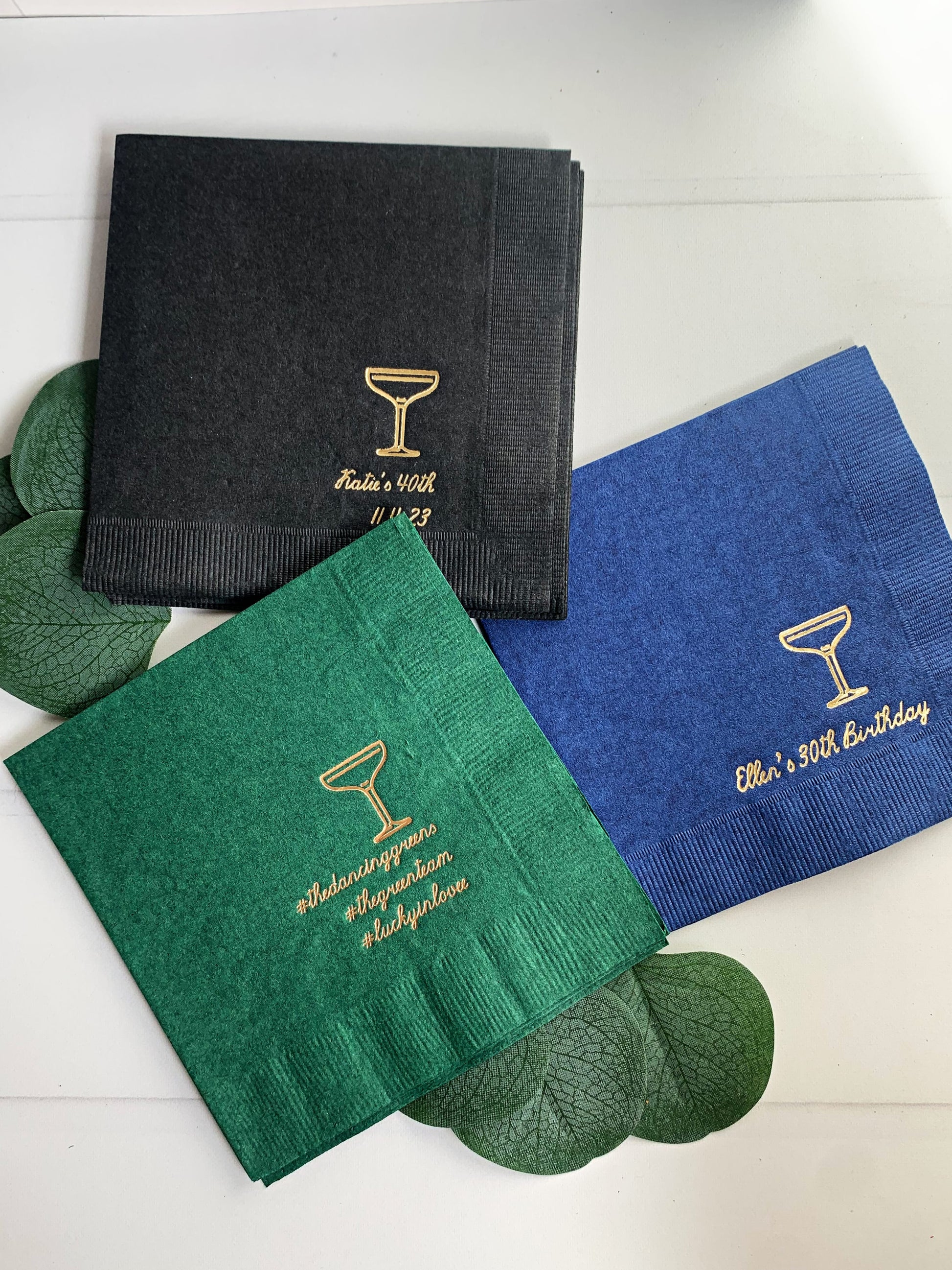Three personalized paper napkins in black, green, and blue, each printed with a gold cocktail glass icon and personalized birthday text, arranged on a white surface with decorative green leaves.