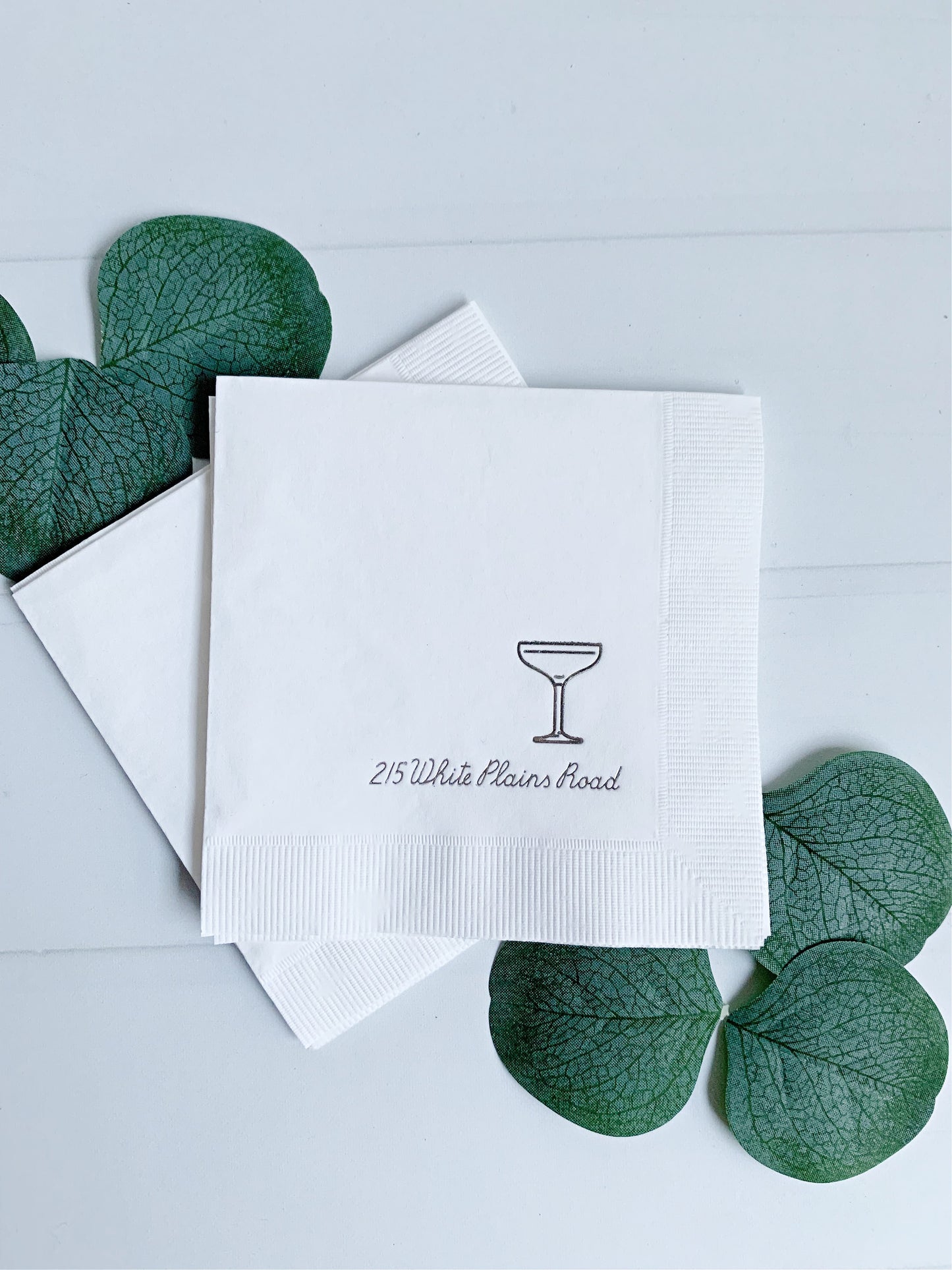 White elegant personalized paper napkins stacked on a light surface, with one napkin printed with a small cocktail glass icon and the text '215 White Plains Road.' Green decorative leaves are placed around the napkins.