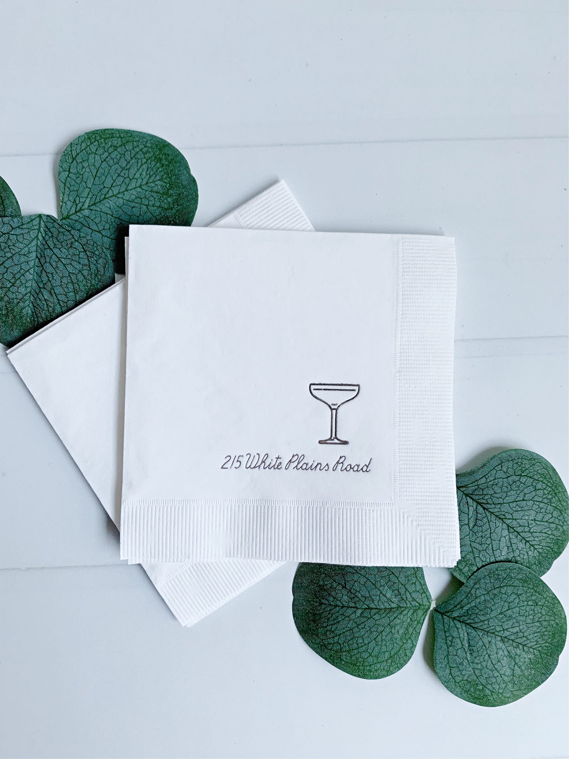 White elegant personalized paper napkins stacked on a light surface, with one napkin printed with a small cocktail glass icon and the text '215 White Plains Road.' Green decorative leaves are placed around the napkins.
