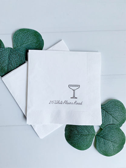 White elegant personalized paper napkins stacked on a light surface, with one napkin printed with a small cocktail glass icon and the text '215 White Plains Road.' Green decorative leaves are placed around the napkins.