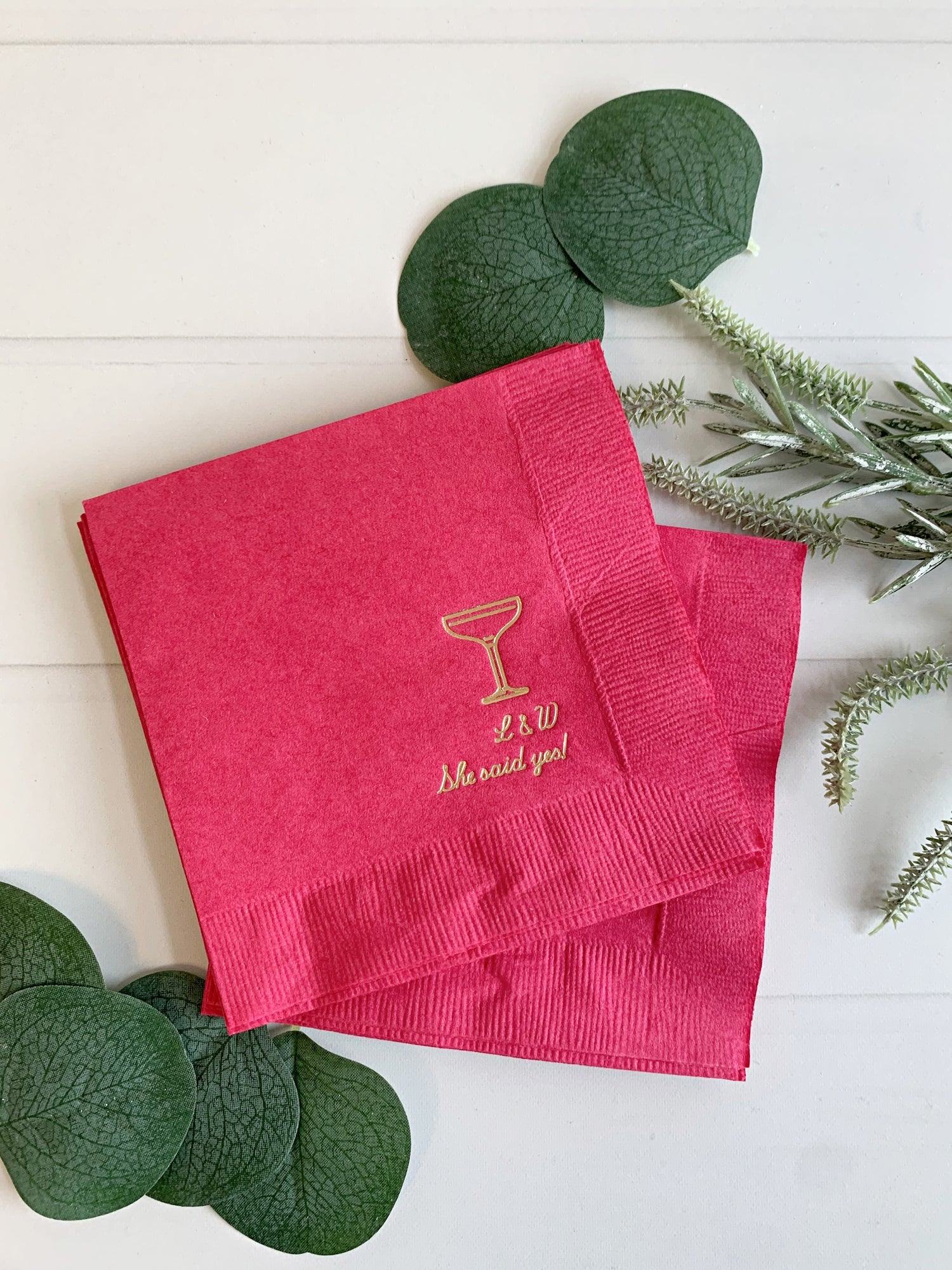 Pink napkin custom engagement party napkin with gold cocktail graphic and text on a white surface with green leaves. Text has initials with 'she said yes!" below.  An EnFete Party original design, copyright 2025.