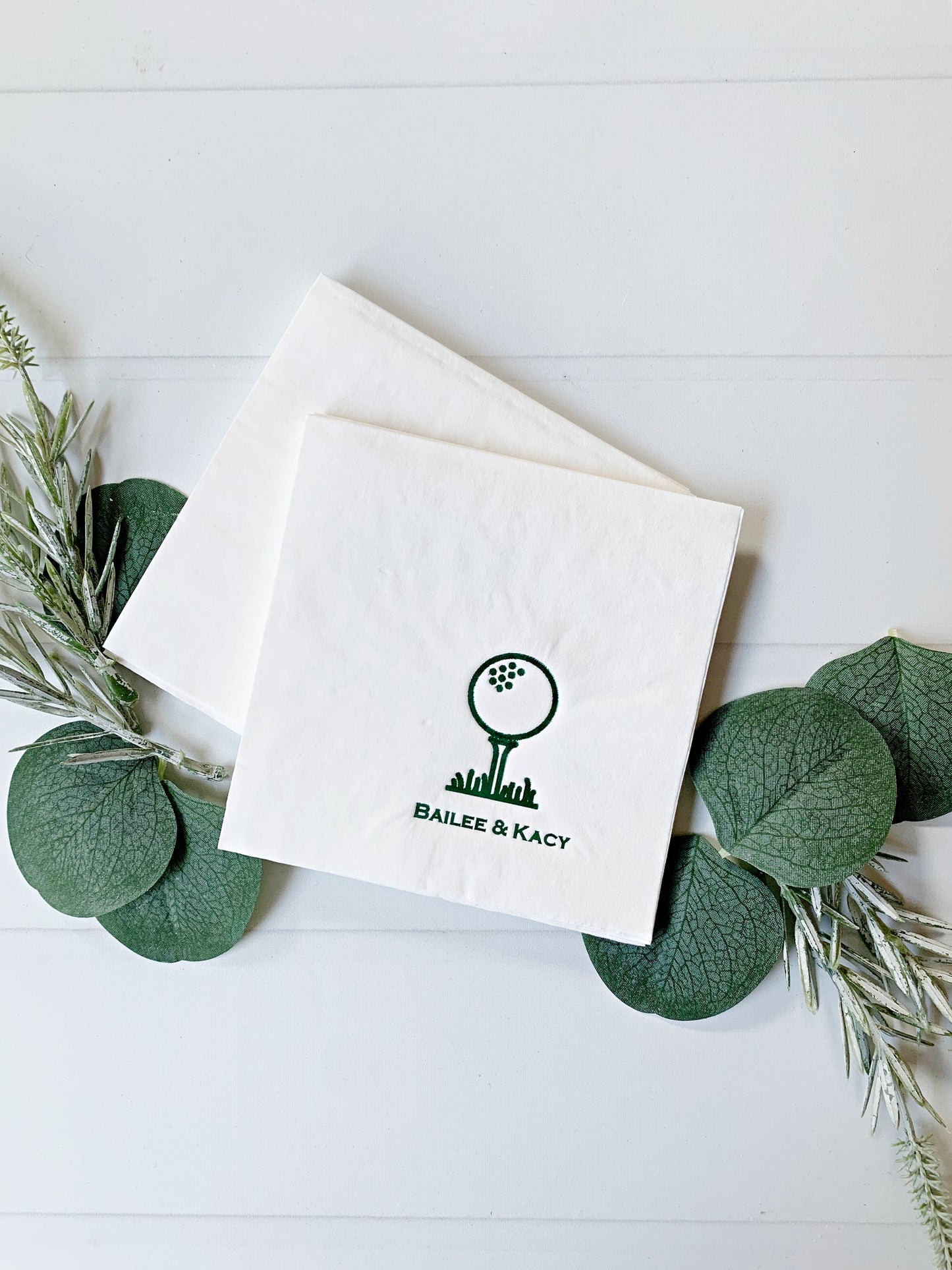 Two white personalized paper cocktail napkins with a green golf ball and club graphic and the text 'BAILEE & KACY' printed on them, arranged on a white wood background with green eucalyptus leaves and sprigs.