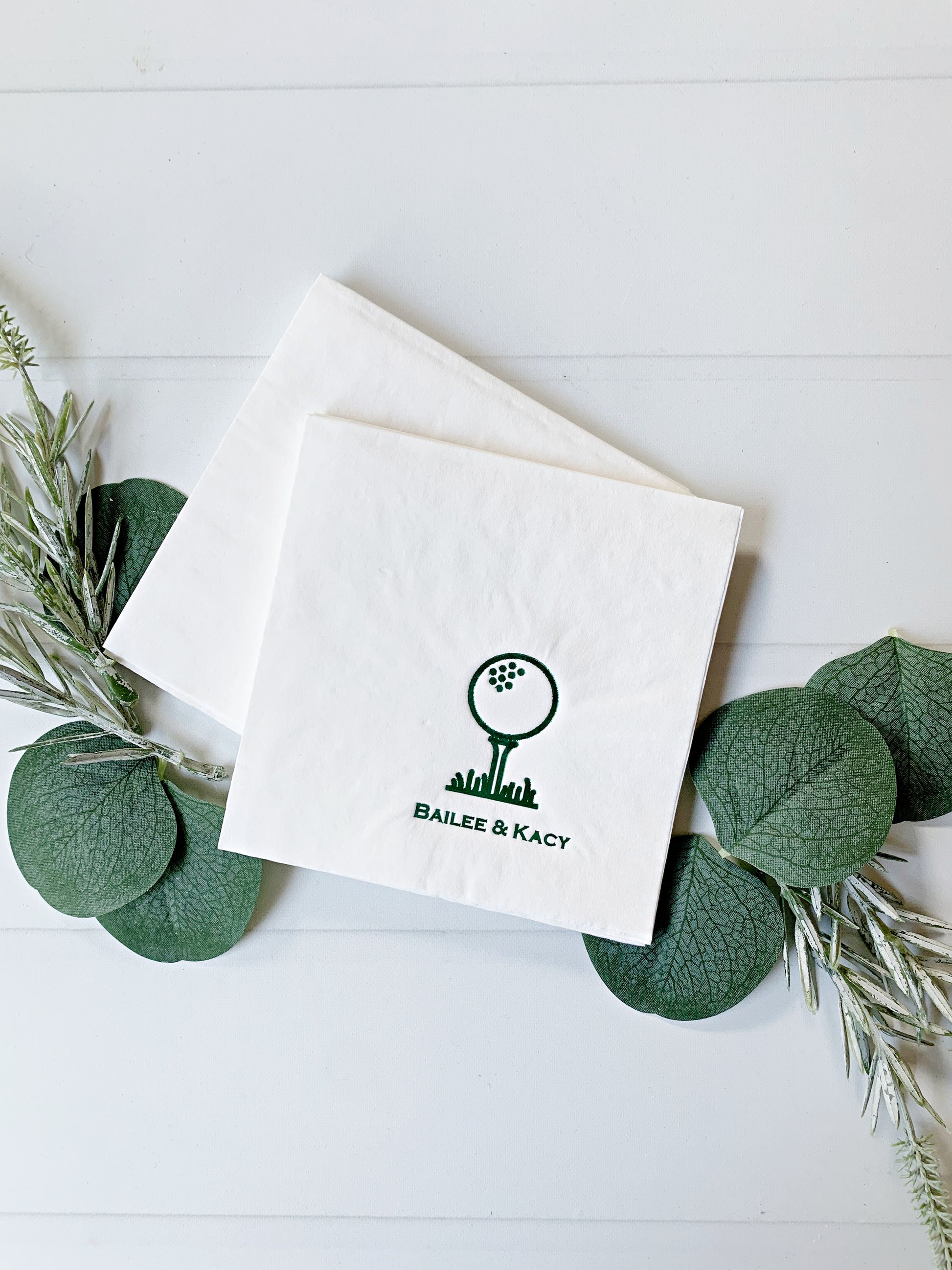 Two white personalized paper cocktail napkins with a green golf ball and club graphic and the text 'BAILEE & KACY' printed on them, arranged on a white wood background with green eucalyptus leaves and sprigs.