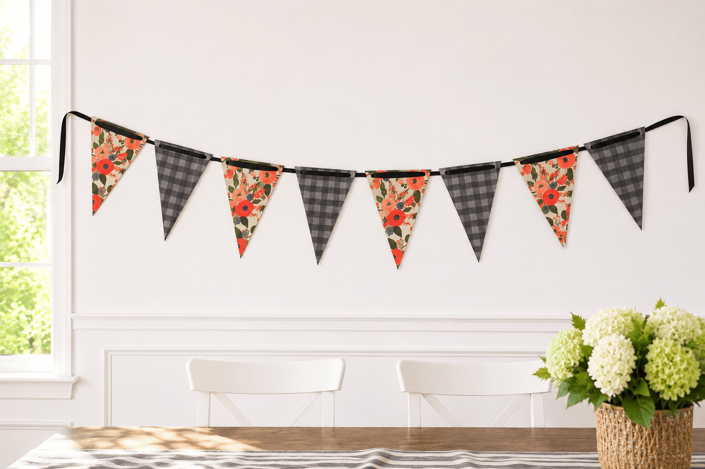 A string of triangular bunting flags hangs on a white wall in a bright room. The flags alternate between charcoal and grey plaid and floral prints with red, orange, and green tones reminiscent of Rifle Paper.