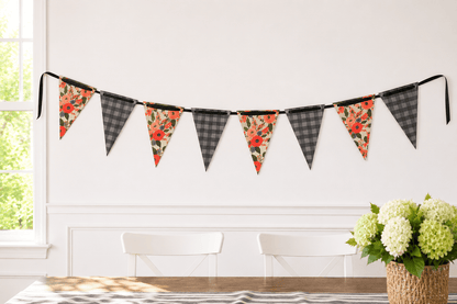 A string of triangular bunting flags hangs on a white wall in a bright room. The flags alternate between charcoal and grey plaid and floral prints with red, orange, and green tones reminiscent of Rifle Paper.
