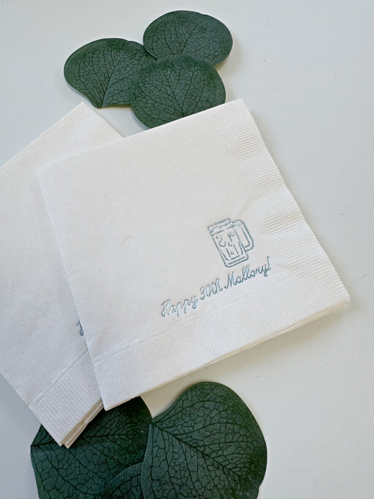 Two white paper napkins stacked on a light surface, with one napkin printed in blue script reading 'Happy 20th Mallory!' next to a blue beer mug graphic. Three dark green heart-shaped decorative leaves are placed around the napkins.