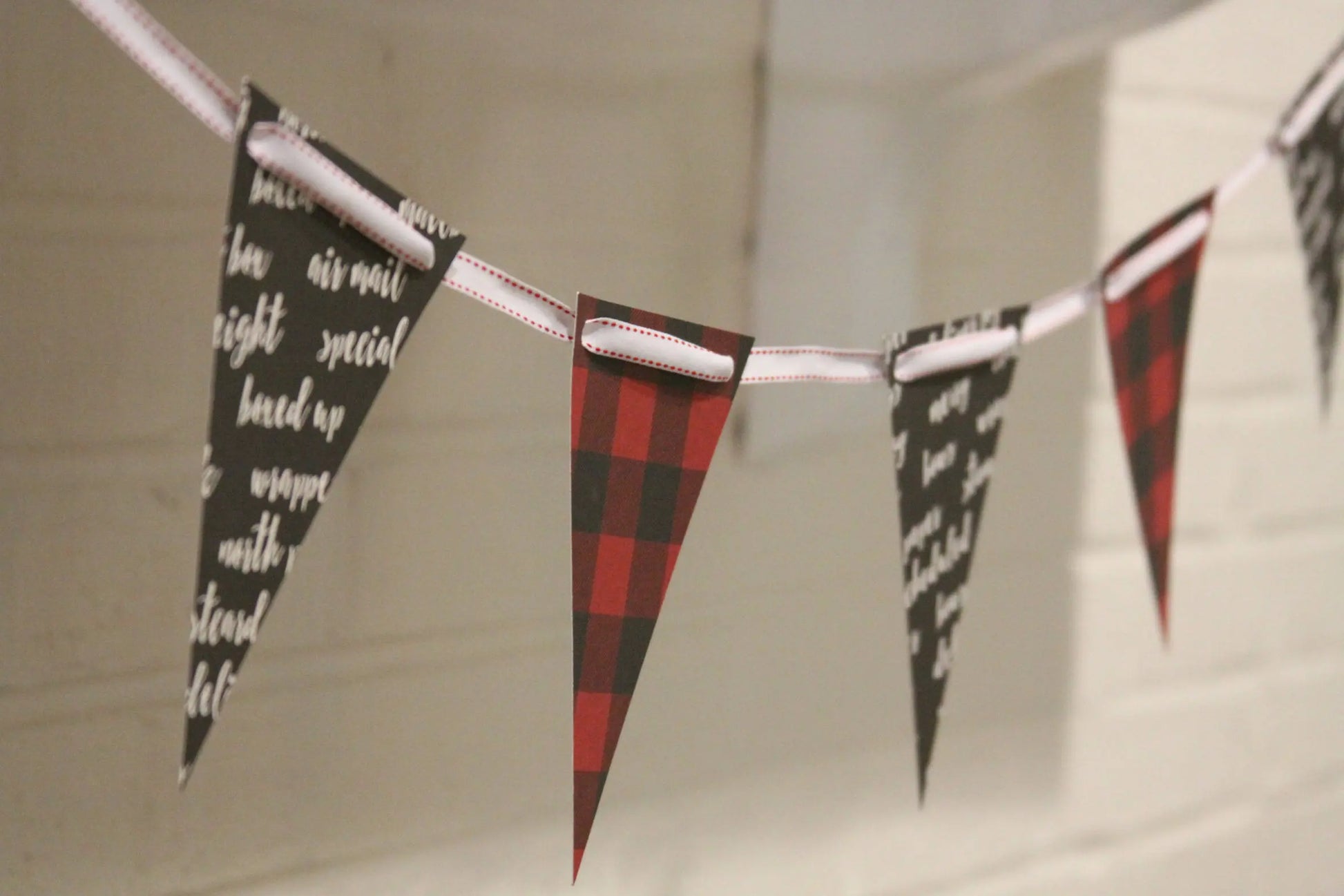 A string of triangular bunting flags hanging across a white brick wall. The flags alternate between black with white script text and red-and-black buffalo plaid.
