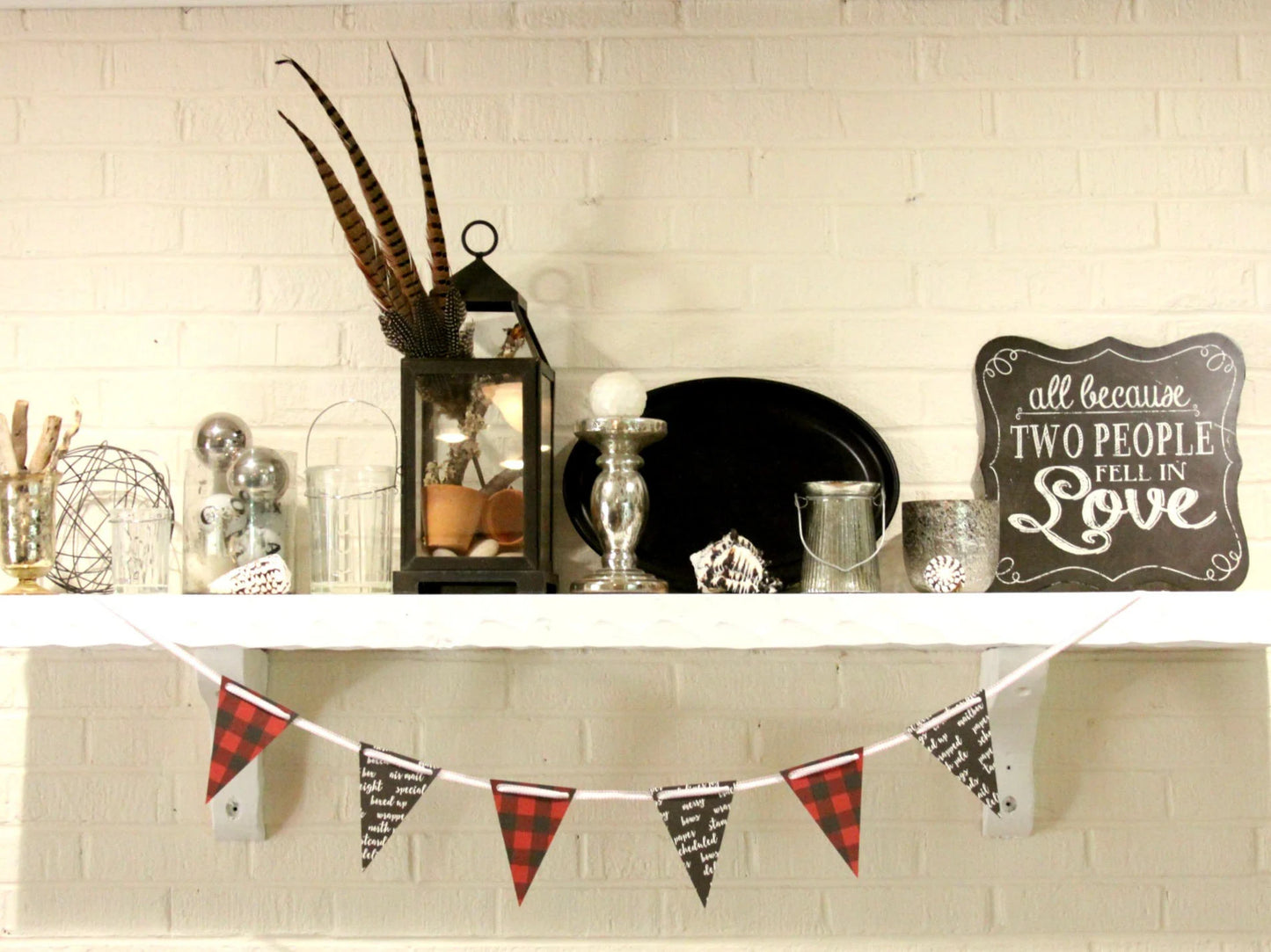A styled holiday mantel display featuring a red and black buffalo plaid bunting banner hanging below a white shelf against a white brick wall. The shelf is decorated with a black lantern with feathers, silver and glass ornaments, a black plate, and a chalkboard sign that reads 'all because TWO PEOPLE LOVE IN Love'.