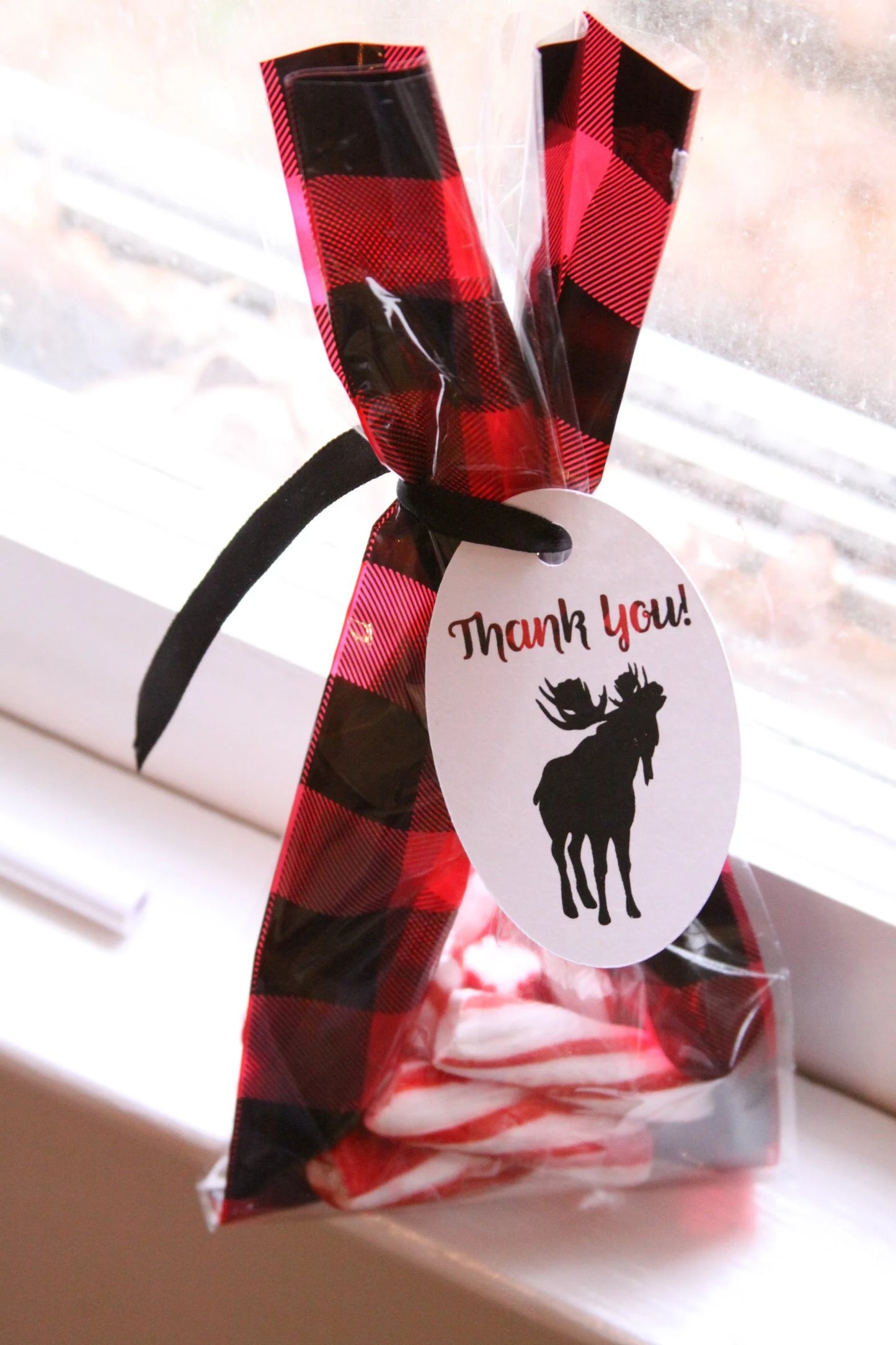 A clear plastic candy bag tied with a red and black buffalo plaid ribbon and black ribbon tail, with a white oval tag that says 'Thank you!' and a black moose silhouette. Candy canes are visible inside, and the bag is photographed on a windowsill with a blurred window background.