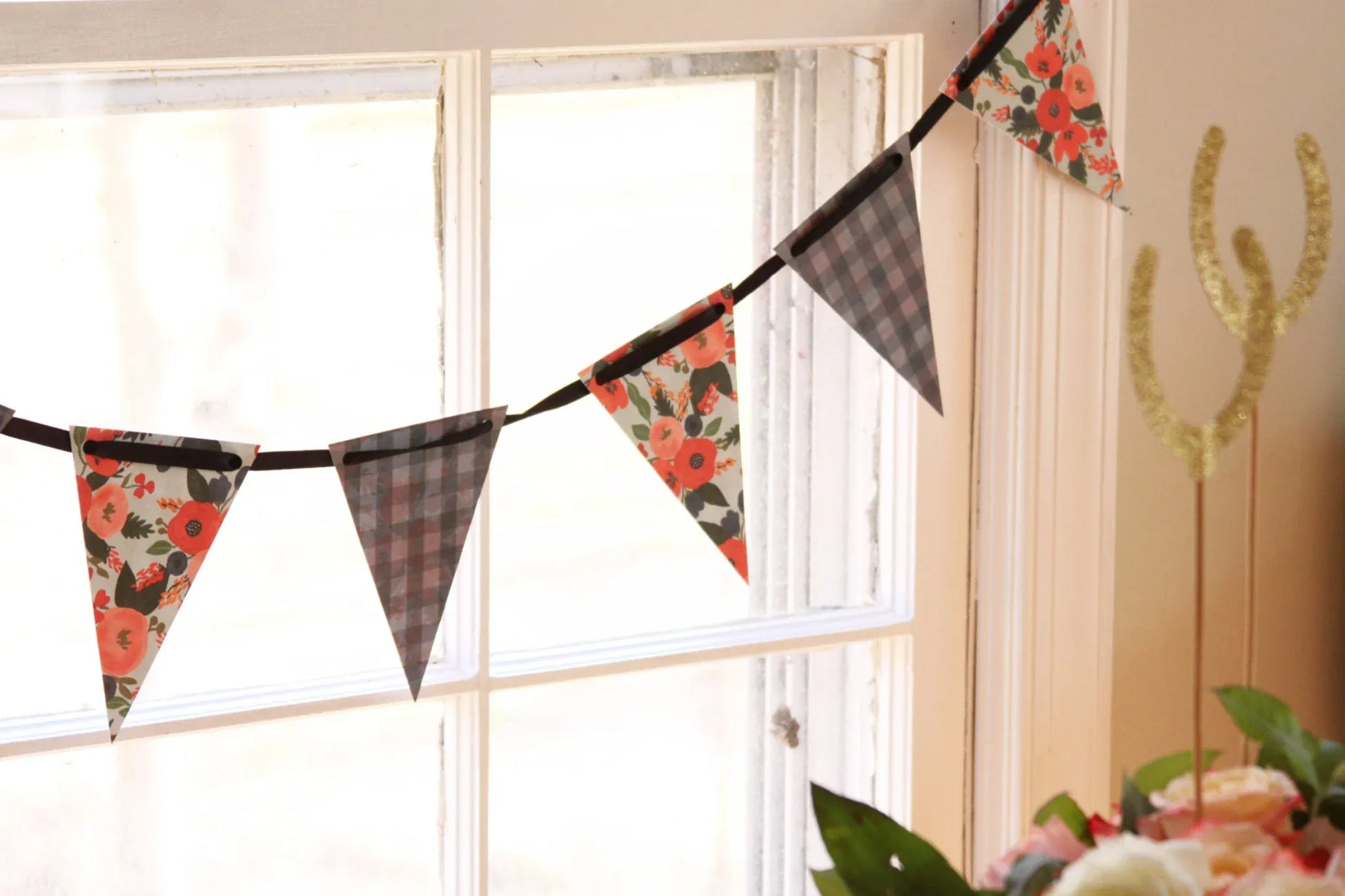 A rustic bunting banner garland with alternating chatcoal grey plaid and floral patterned triangular flags, hanging in front of a bright window in a home setting.