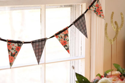 A rustic bunting banner garland with alternating chatcoal grey plaid and floral patterned triangular flags, hanging in front of a bright window in a home setting.