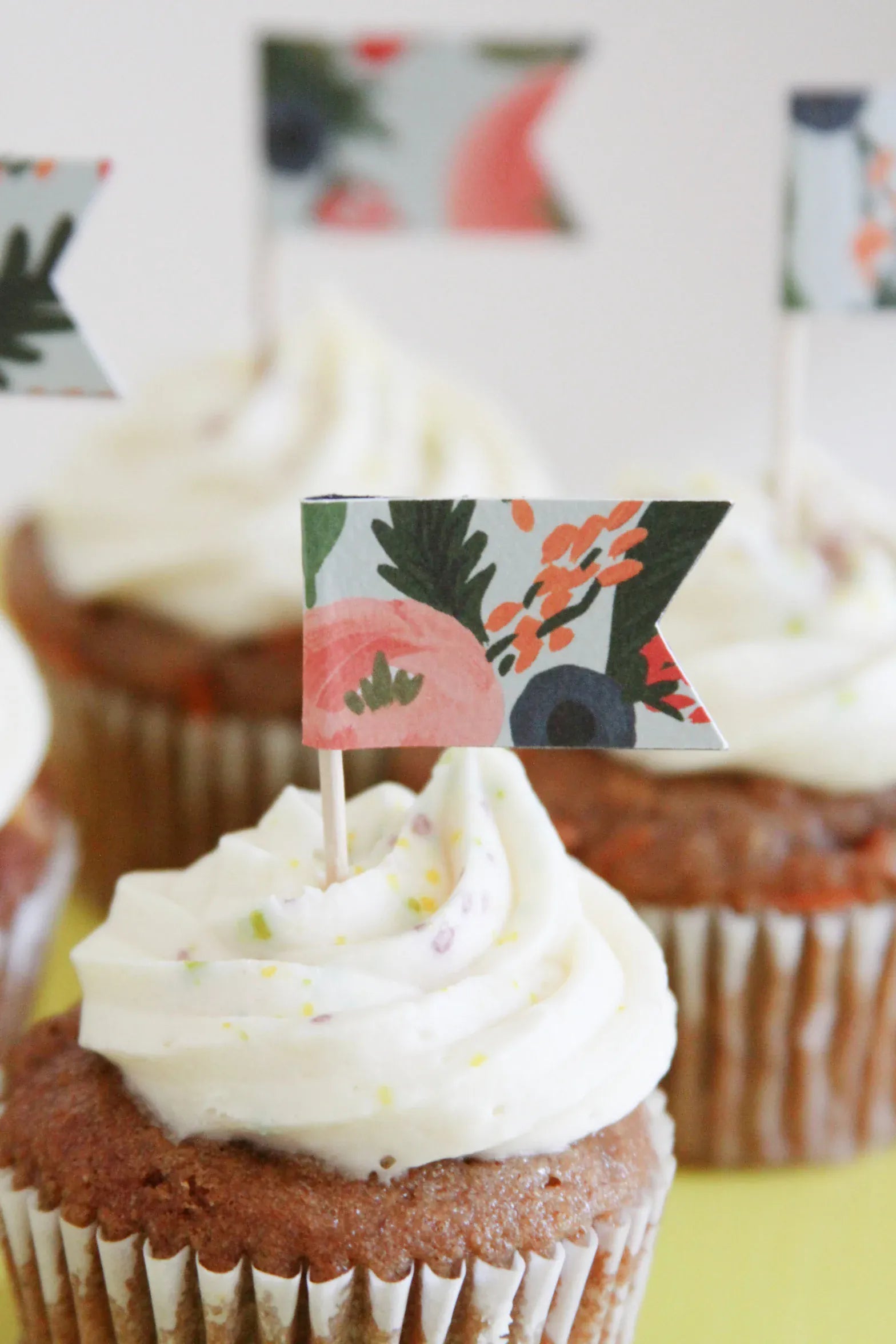 A close-up of a frosted cupcake with a small decorative floral coral and poppy paper topper flag on a toothpick, with additional similar cupcakes blurred in the background.