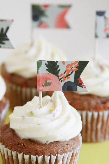 A close-up of a frosted cupcake with a small decorative floral coral and poppy paper topper flag on a toothpick, with additional similar cupcakes blurred in the background.