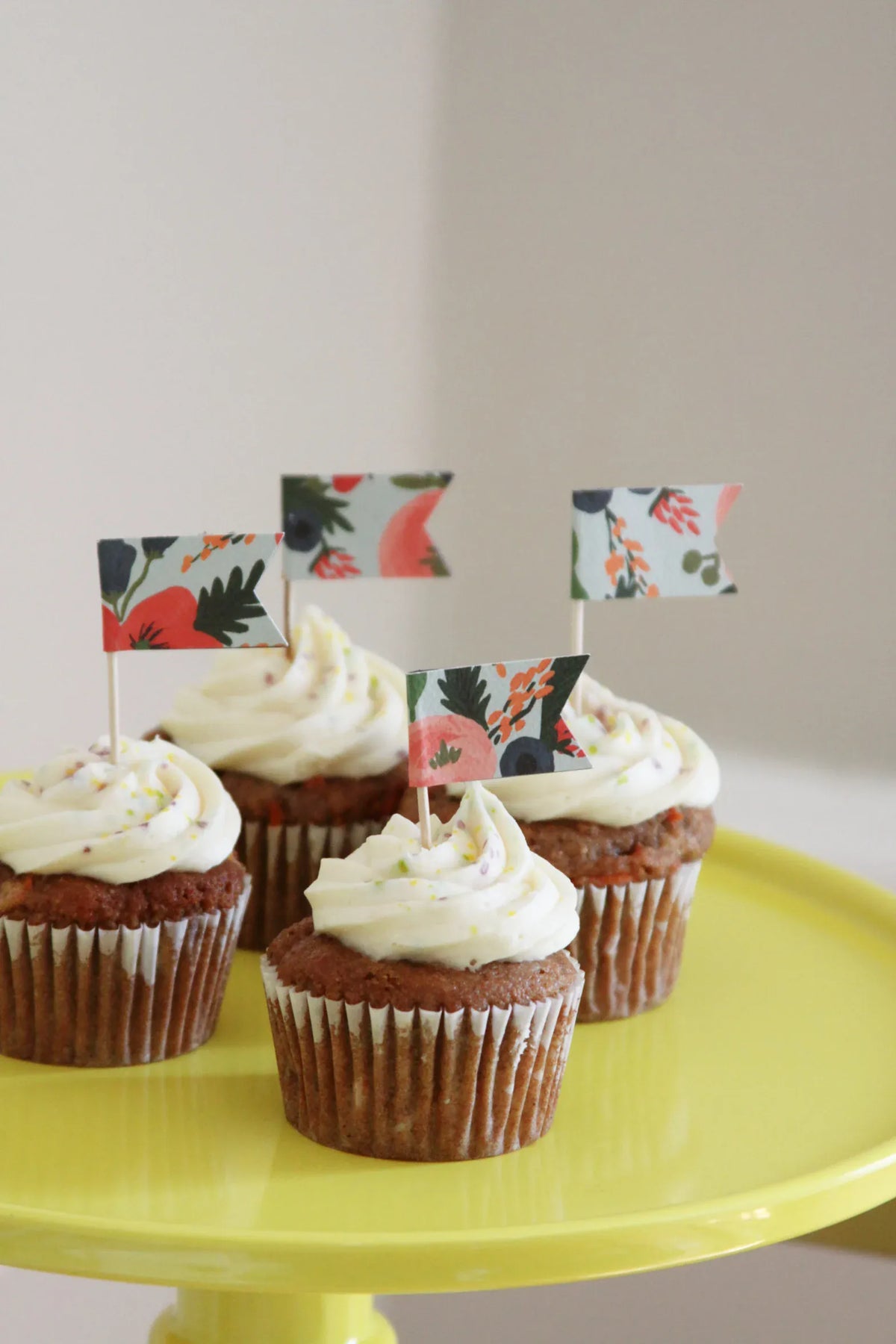 Four frosted cupcakes with small decorative coral pink and blue floral picket flags on a bright yellow cake stand against a soft beige background ready for baby in bloom shower decorations.  Handmade by EnFete Party.