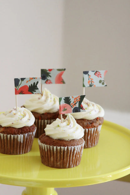Four frosted cupcakes with small decorative coral pink and blue floral picket flags on a bright yellow cake stand against a soft beige background ready for baby in bloom shower decorations.  Handmade by EnFete Party.