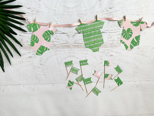 A tropical-themed baby shower decoration set featuring three paper baby bodysuit banners in pink and green leaf patterns, hanging from a pink ribbon with wooden clothespins, along with matching paper cupcake toppers on toothpicks, arranged on a white wood background with a green palm leaf prop.