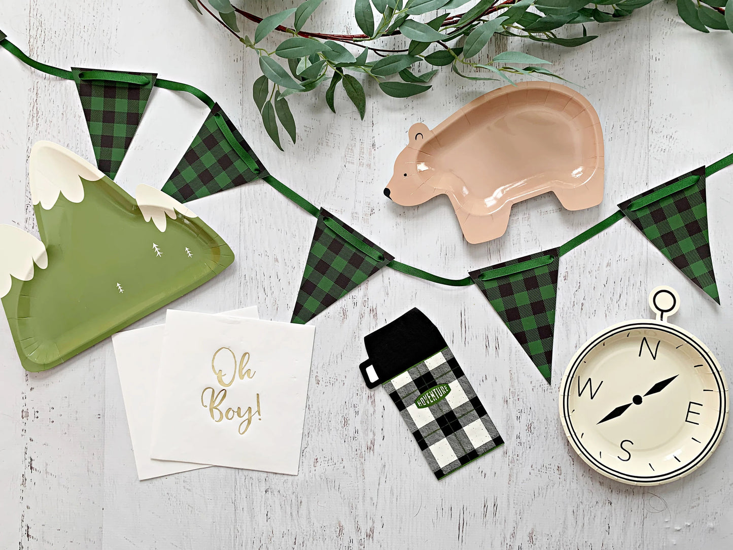 A styled flat-lay of woodland-themed party supplies on a white wood background, including green and black plaid bunting, green mountain-shaped paper plates, a bear-shaped paper plate, napkins with gold foil text, a small plaid pouch, and a compass.